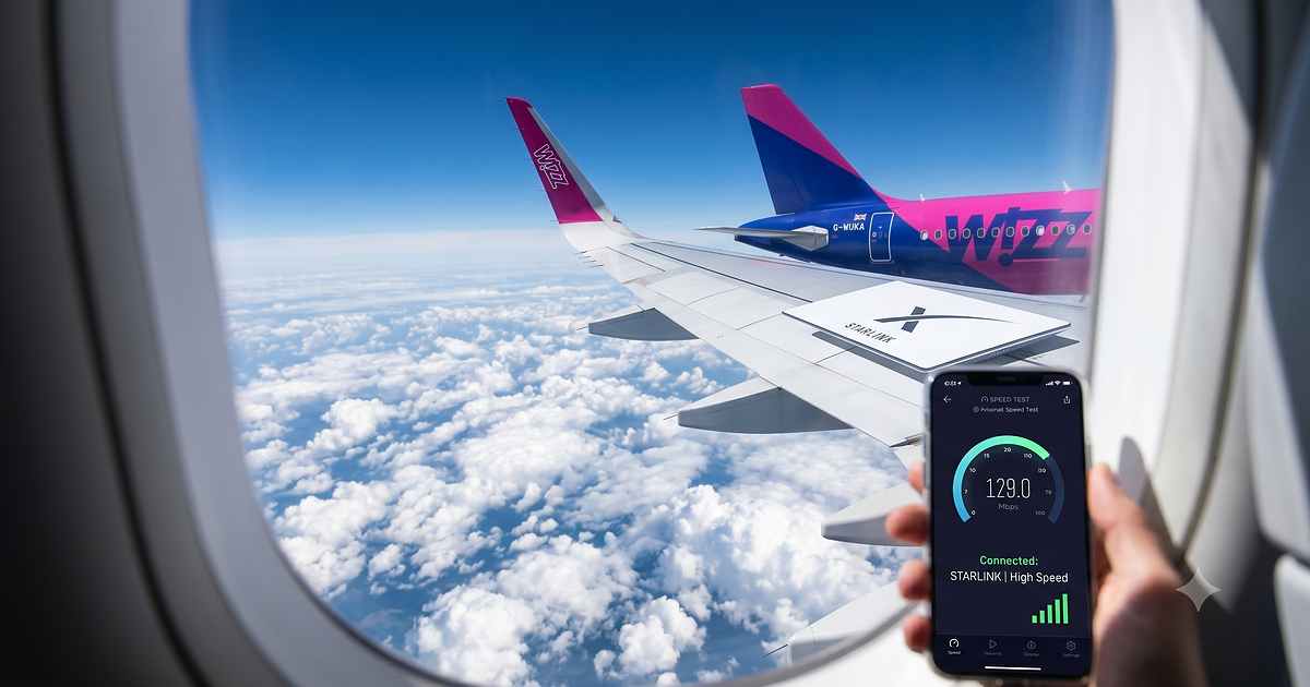 Pogled skozi okno letala Wizz Air na oblake, kjer potnik na telefonu izvaja test hitrosti povezave Starlink.
